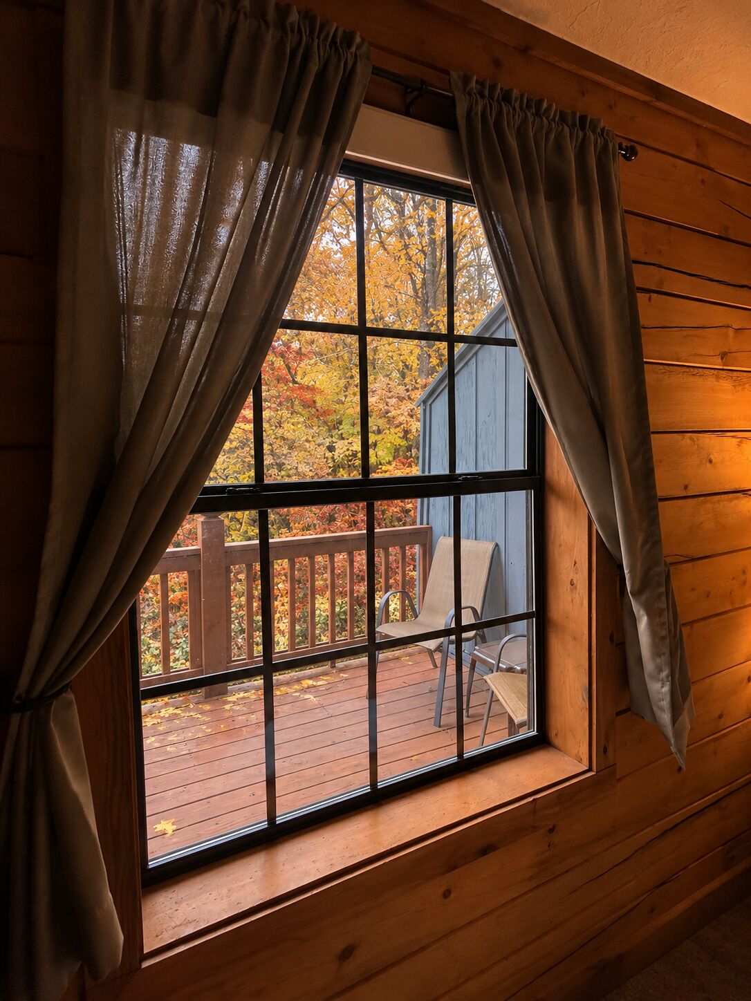 A window with curtains, a wooden deck, and a chair.
