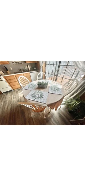 A round dining table with a floral tablecloth, surrounded by white chairs, in a room with wooden flooring and large windows.