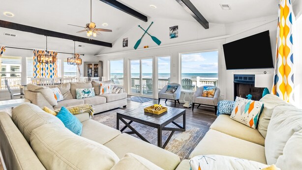 A spacious living room with a large sofa, a coffee table, and a TV mounted on the wall. There are large windows offering a view of the ocean.