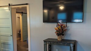 A room with a large flat-screen TV mounted on the wall, a rustic wooden side table with a plant, and a door leading to another room.