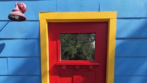 A red door with a yellow frame, a red mailbox, and a colorful doormat with "HELLO" on a concrete floor.