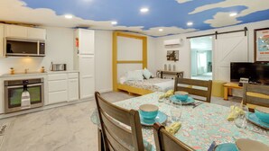 Una camera compatta con angolo cottura, zona pranzo e un letto. La stanza presenta un soffitto con decoro a nuvole, mobili bianchi e un letto con struttura in legno.