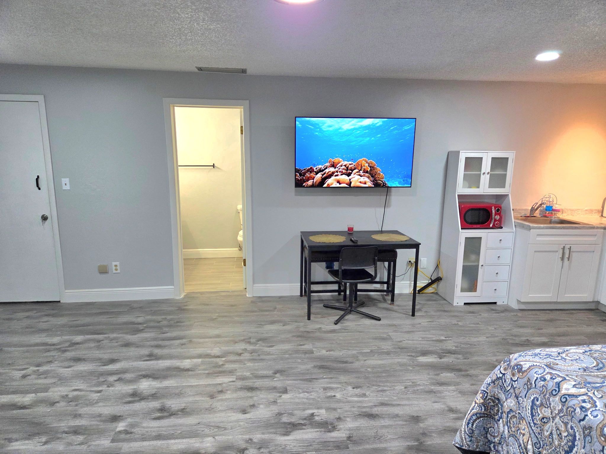A bedroom with a bed, a TV mounted on the wall, a desk with a chair, and a kitchenette area.