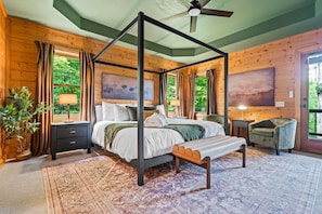 A four-poster bed with a bench, a bedside table, a lamp, and a view of greenery outside.