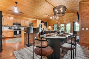 A wooden cabin interior with a kitchen, dining area, and a large window.