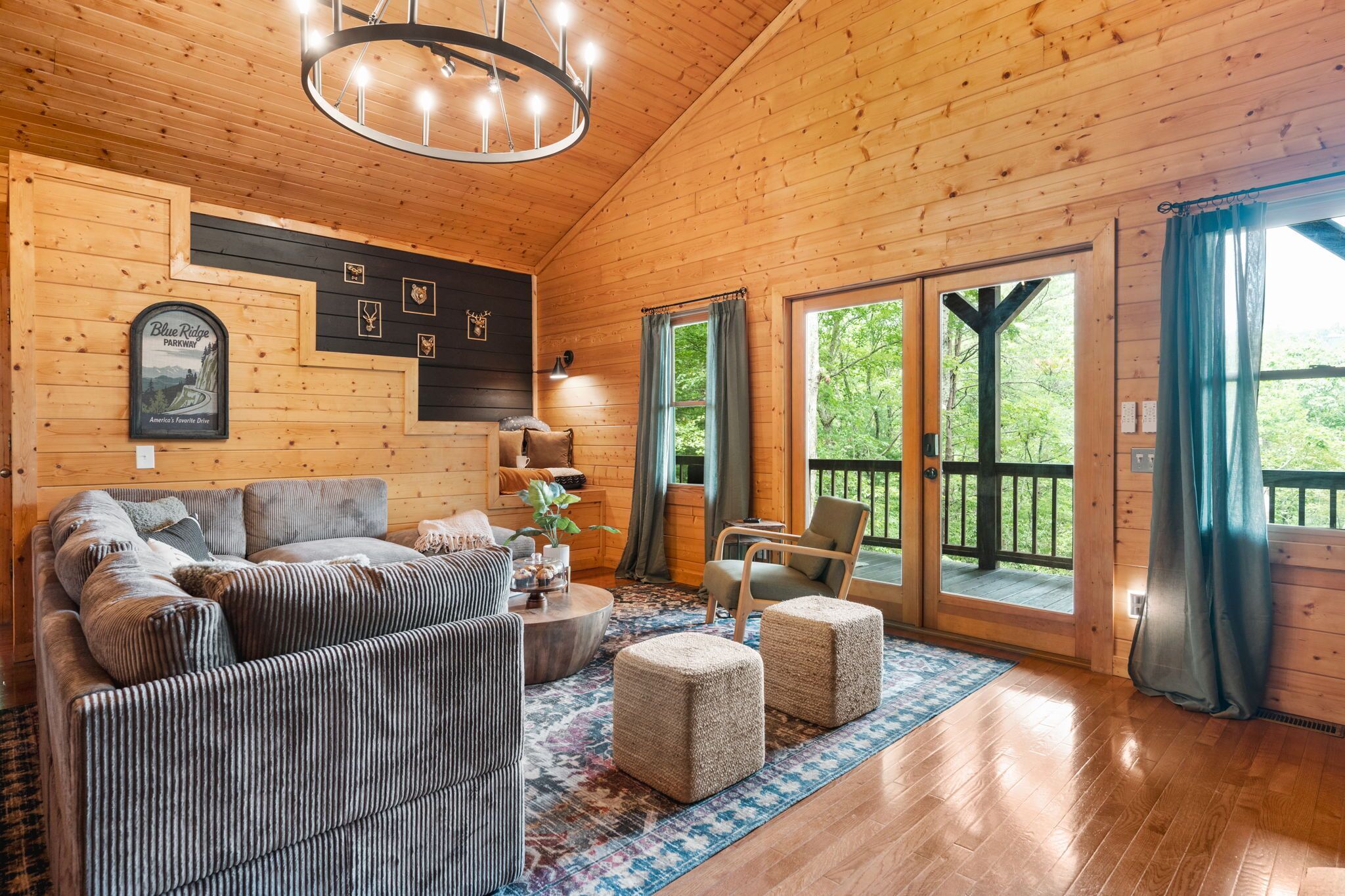 A wooden cabin interior with a sofa set, ottomans, a chandelier, and a large window with a view of greenery.