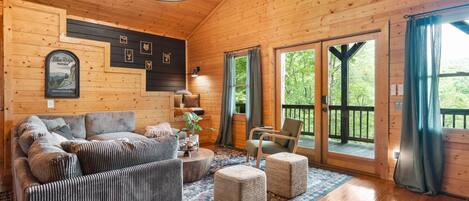 Ein Holzblockhaus-Innenraum mit einer Sofagarnitur, Poufs, einer Kristallleuchter und einem großen Fenster mit Blick auf Grünflächen.