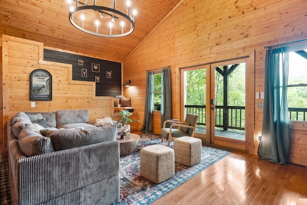 A wooden cabin interior with a sofa set, ottomans, a chandelier, and a large window with a view of greenery.