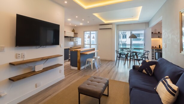 Una sala moderna con televisor de pantalla plana, estanterías de madera, un sofá oscuro y un comedor con mesa y sillas.