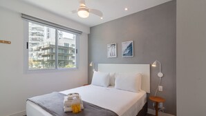Una habitación de hotel moderna con una cama grande, un ventanal con vistas a la ciudad, un ventilador de techo y una mesita de noche con lámpara.