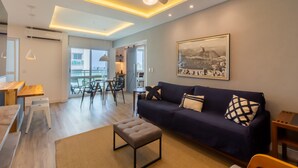 Una sala moderna con un sofá azul oscuro, un otoman beige y un comedor con muebles de madera.