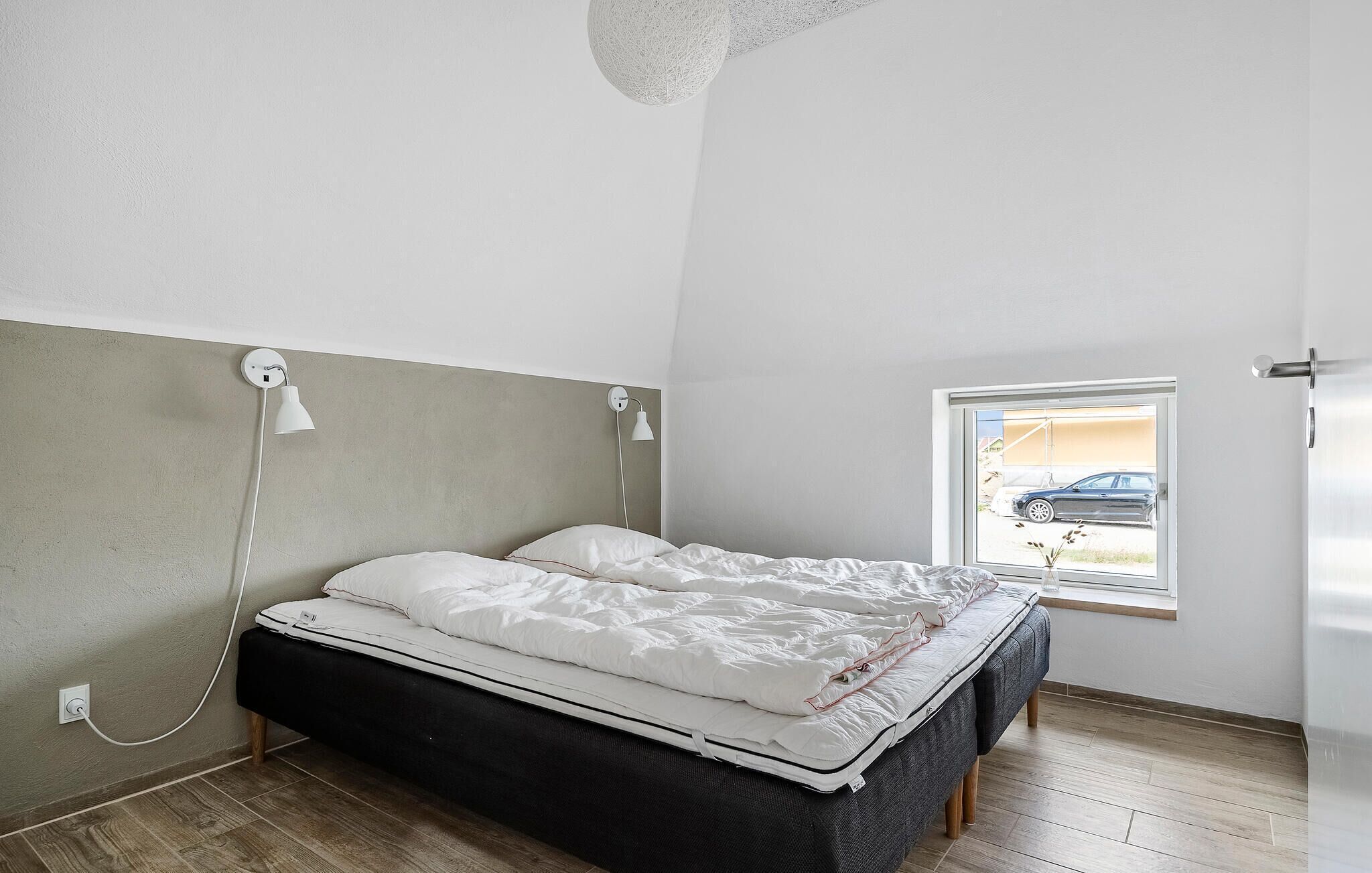 A bedroom with a bed, a window with a view of a car, and a wall-mounted lamp.