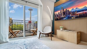 Quarto com uma janela grande, uma cadeira, uma mesa pequena, um armário e um quadro de uma paisagem urbana.