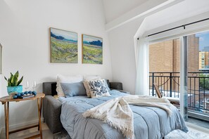 Une chambre à coucher avec un lit, une petite table sur laquelle se trouvent une plante et des verres, deux tableaux accrochés au mur, et une vue sur la ville depuis un balcon.