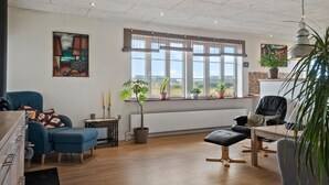 Un salon moderne avec un canapé bleu, une table à manger en bois et un fauteuil en cuir noir. Plusieurs plantes en pot sont disposées sur le rebord de la fenêtre, et un tableau orne le mur.