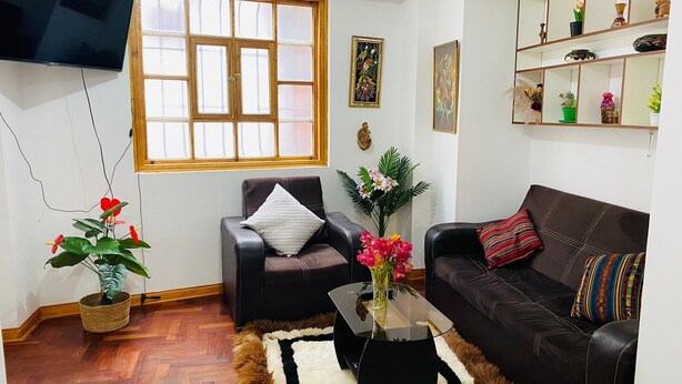 A living room with a wooden floor, a black leather sofa, a black leather armchair, a glass coffee table, and a white rug. - Relax in San Blas - 3-Bedroom Apartment with Views 🌅 (Cusco)