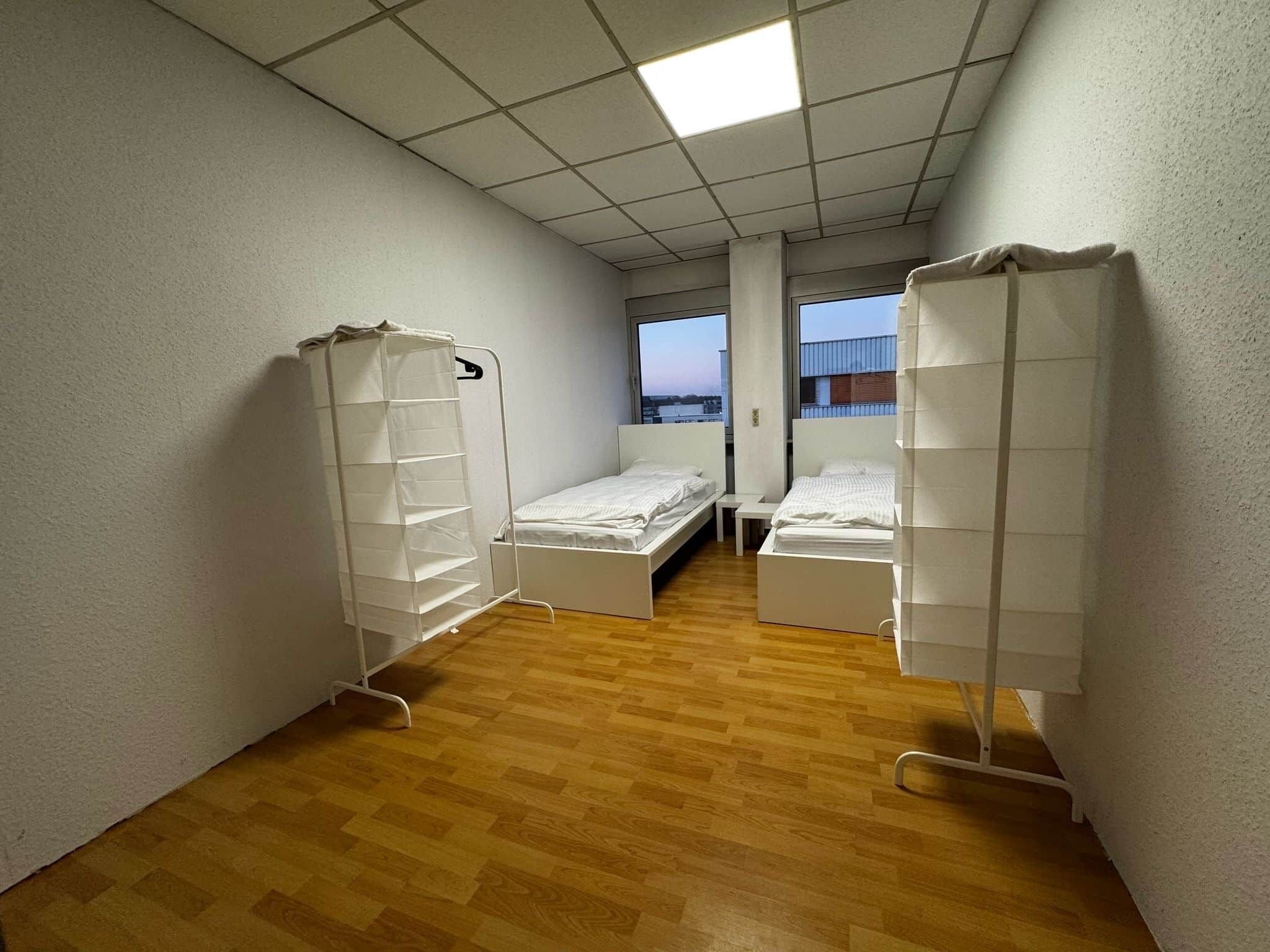 Quarto com duas camas, uma janela e vista para um edifício.