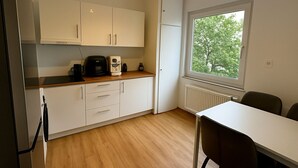 A compact kitchen with white cabinets, a coffee maker, and a radiator near a window with a view of trees. - Mainz 3 Betten mit Parkplatz (Mainz)