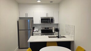 A compact kitchen with white cabinets, a stainless steel refrigerator, a microwave, and a sink. There is a small island with a white countertop and two yellow chairs. - Cozy retreat (Edmonton)