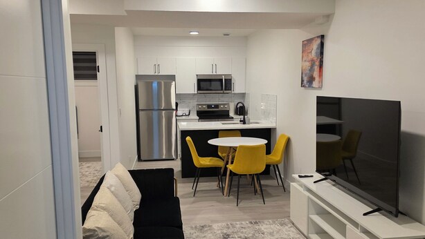 A modern living space with a sofa, a dining area with yellow chairs, a kitchen with a refrigerator and microwave, and a TV on a stand. - Cozy retreat (Edmonton)