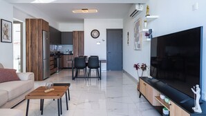 Una sala de estar moderna con sofá, mesa de centro, televisor y un comedor con sillas, con una cocina al fondo.