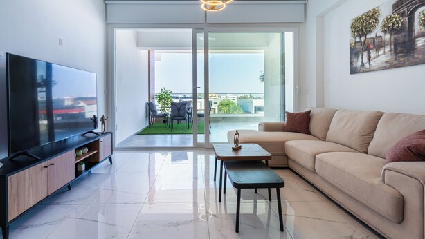 Una sala de estar moderna con una televisión de pantalla plana, un sofá beige, una mesa de centro y un comedor con sillas y una pequeña mesa.