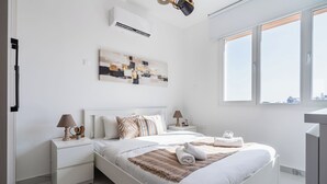 Un dormitorio moderno con una cama grande, dos mesitas de noche con lámparas, aire acondicionado en la pared y vistas a la ciudad desde la ventana.