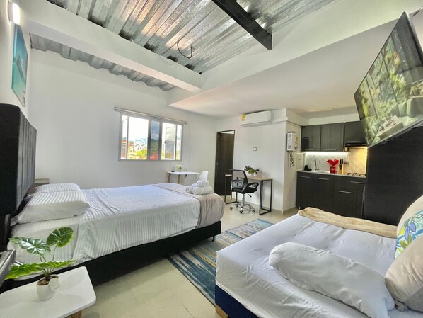 A compact hotel room with two beds, a small kitchenette, a desk, and a window with a city view.