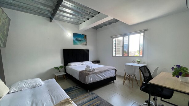A hotel room with a bed, a desk, a chair, a small table, a potted plant, and a painting on the wall. - Edificio RG33 (Medellín)