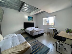 A hotel room with a bed, a desk, a chair, a small table, a potted plant, and a painting on the wall.