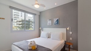 A modern hotel room with a large bed, a window with a city view, a ceiling fan, and a bedside table with a lamp. - Charming Barra 1min From the Beach Lc201b (Rio de Janeiro)