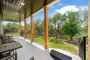 A covered outdoor patio with wooden columns, a black metal railing, and a view of a grassy area with trees.