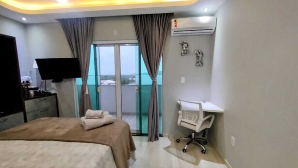 A hotel room with a bed, a desk, a chair, an air conditioner, and a view through the glass doors.
