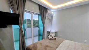 A hotel room with a bed, a TV mounted on the wall, a small table with a chair, and a balcony with curtains.