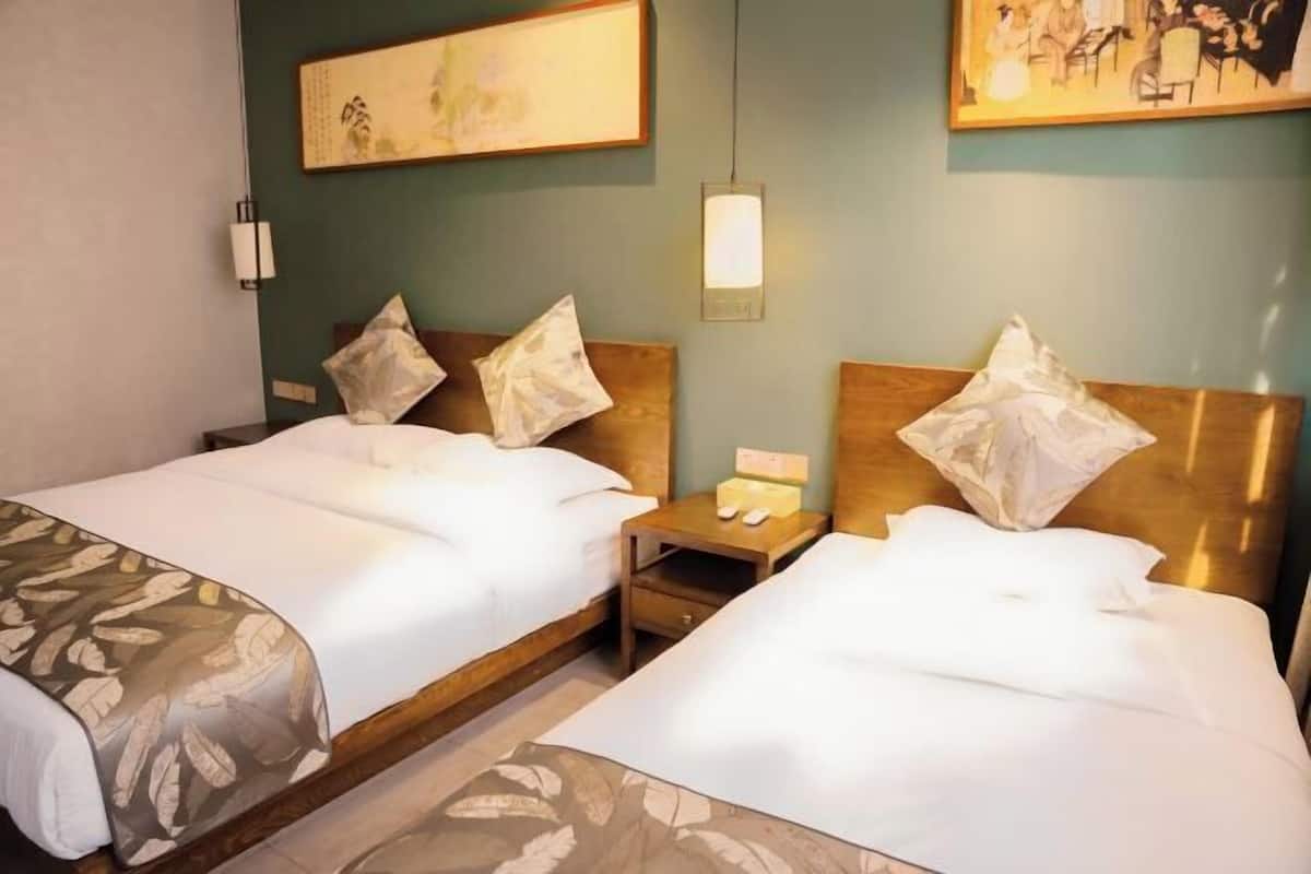 A hotel room with two beds, a nightstand, a wall-mounted lamp, and artwork on the walls.
