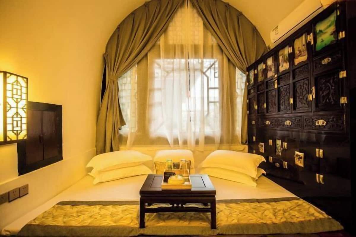 A room with a bed, a bedside table with a tray, a window with curtains, and a dark wooden cabinet with pictures.
