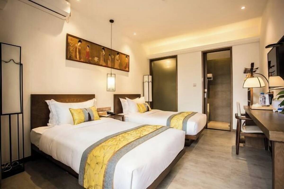 A hotel room with two beds, a painting on the wall, a desk with a lamp, and a door leading to another room.