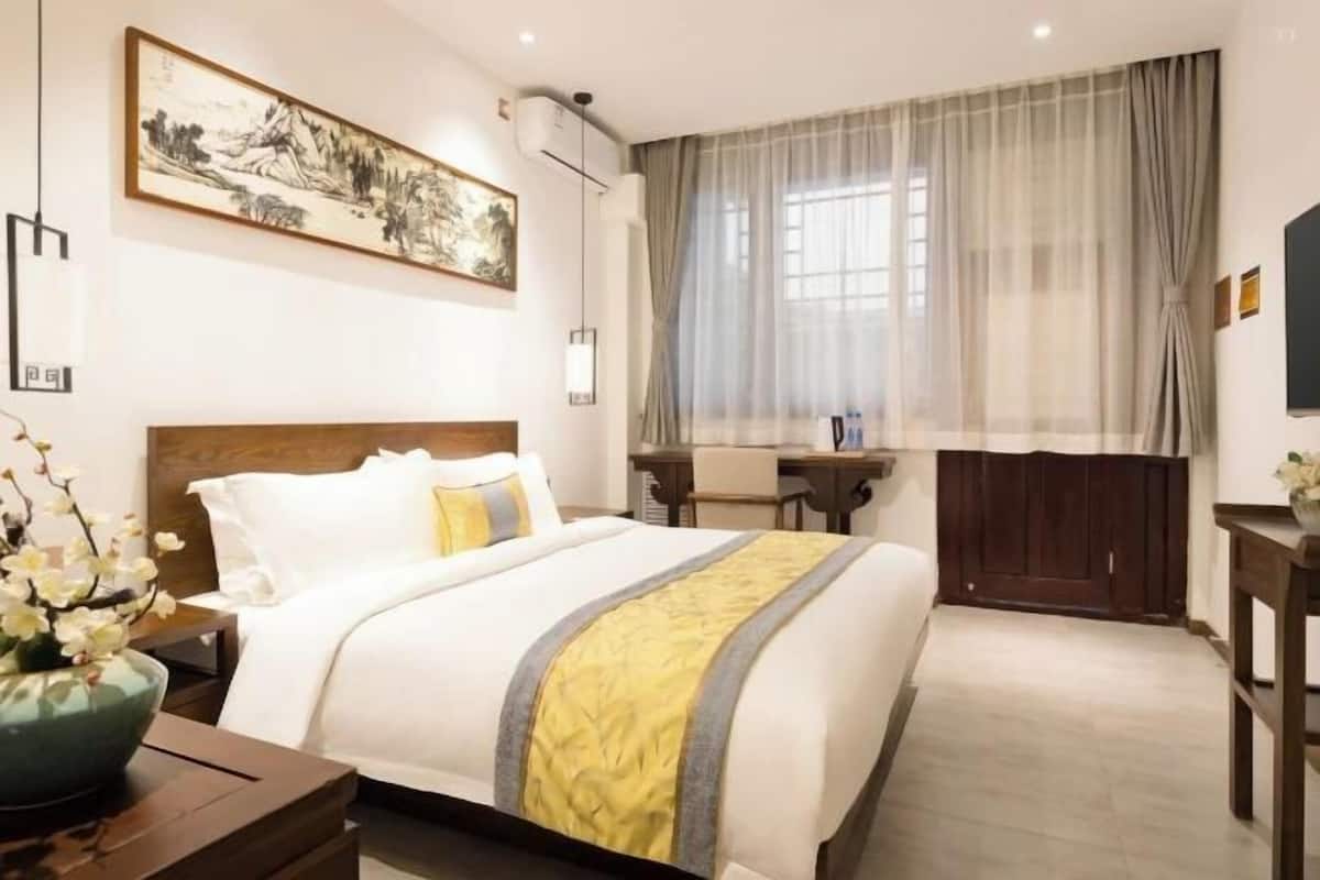 A hotel room with a bed, a desk, a chair, a window with curtains, and a wall-mounted air conditioner.
