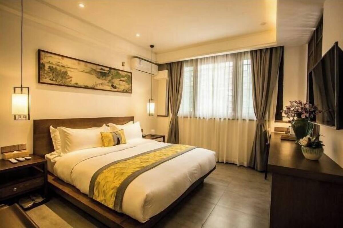 A hotel room with a large bed, bedside tables, a mounted artwork, a window with curtains, and a potted plant.