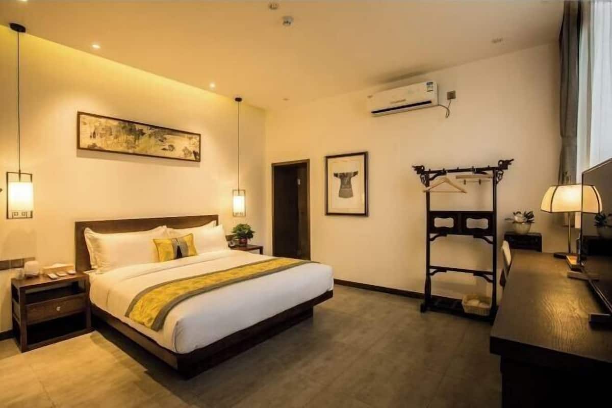 A hotel room with a bed, bedside table, lamp, artwork, and a coat rack.
