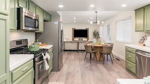Una cocina moderna con armarios verdes, electrodomésticos de acero inoxidable, una zona de comedor con mesa y sillas, y un amplio ventanal con persianas.