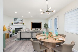 A modern dining area with a round wooden table, beige chairs, and a television mounted above the fireplace. - Remodeled 4BR Home w/Game Room & Backyard Deck (Reno)