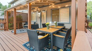 Un patio techado con terraza de madera, mobiliario exterior, luces colgantes y vista de la casa.