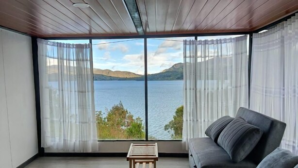 A bedroom with a large bed, a small table, and a view of a lake and mountains through sheer curtains. - SAN ALEJO ECOHOTEL (Tota)