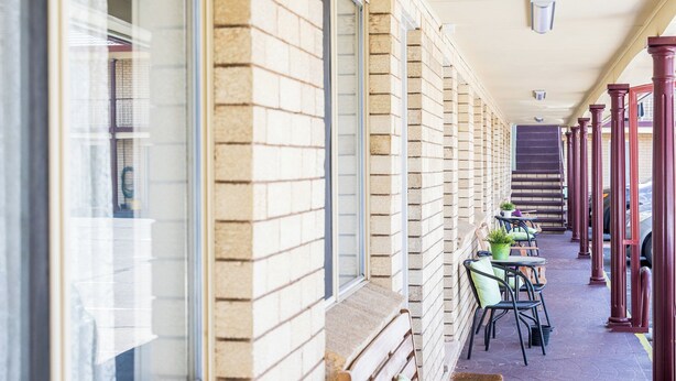 A covered outdoor seating area with wooden benches and chairs, a brick wall, and a window with a view. - Lilac City Motor Inn 2 Bedroom Interconnect 9/10 (Goulburn)