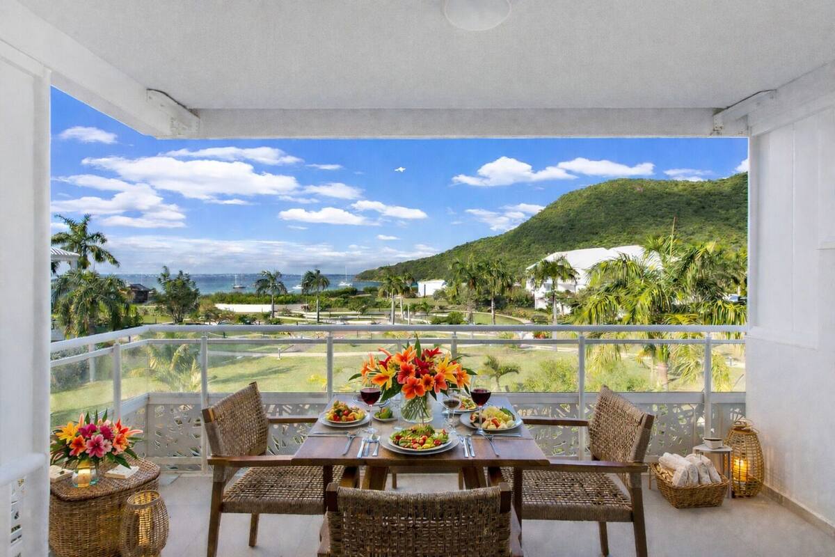 A dining area with a table set for two, overlooking a scenic view of a coastal landscape with palm trees and a mountain.