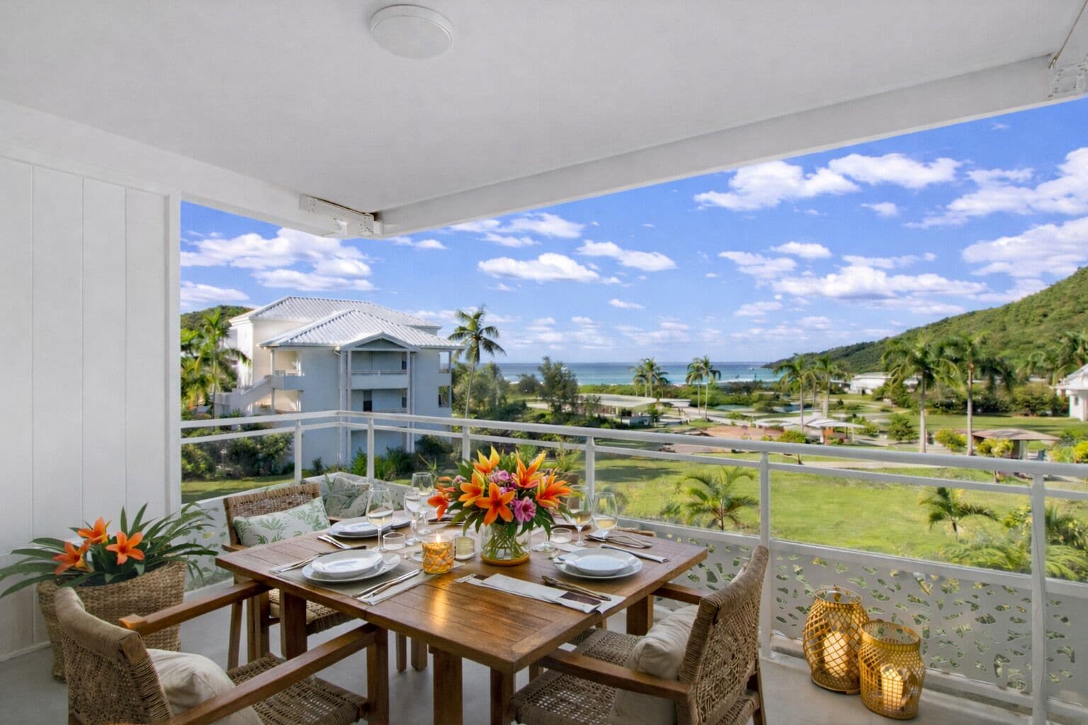 A balcony with a wooden table set for dining, wicker chairs, and a view of a resort with palm trees and a building in the background.