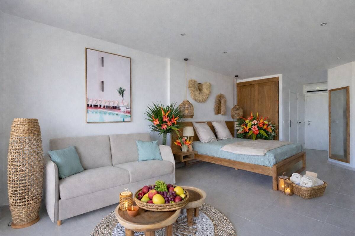 A modern living room with a sofa, a bed, a dining table with fruit, and a large vase of flowers.