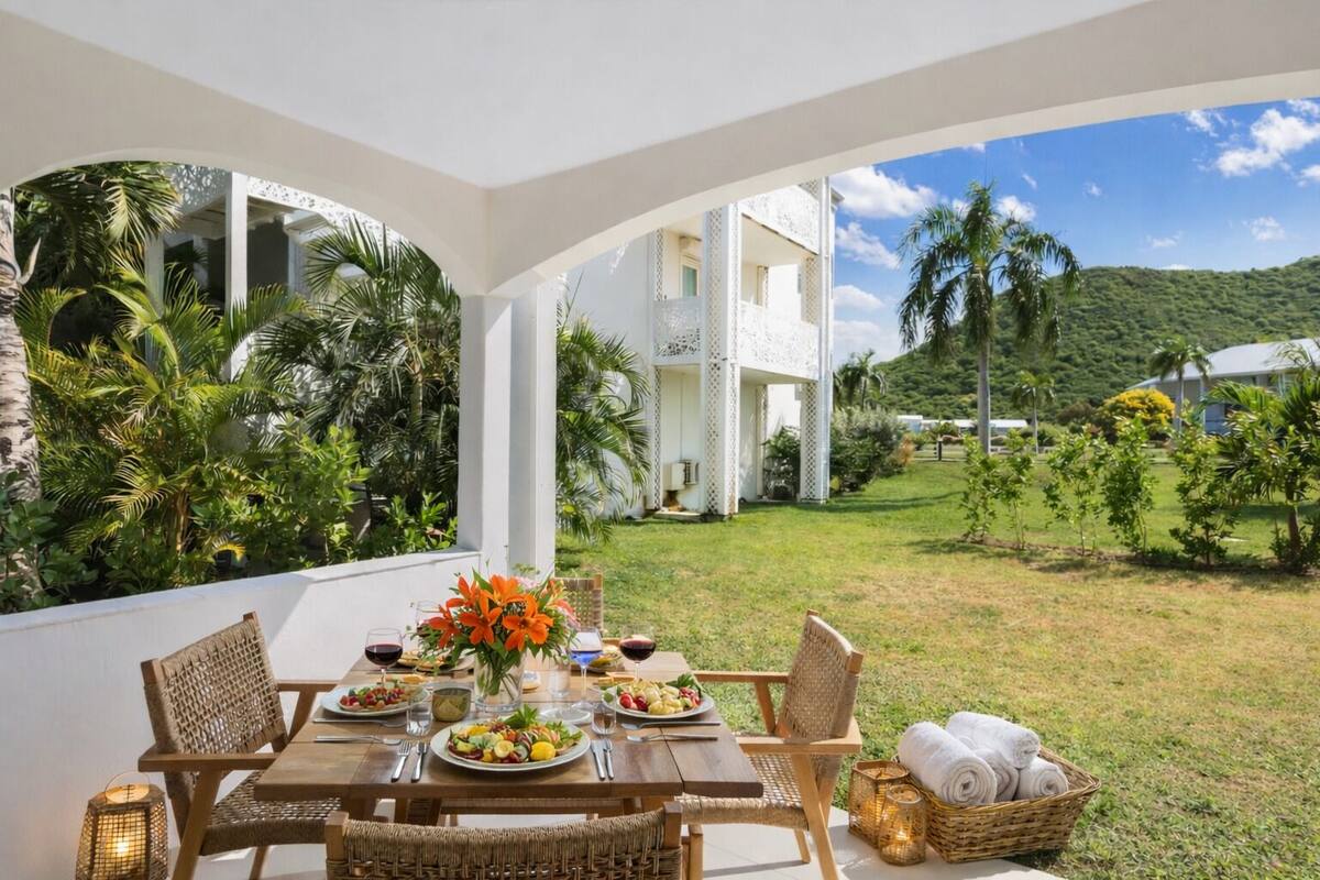 An outdoor dining area with a table set for two, surrounded by lush greenery and a view of a white building with balconies.
