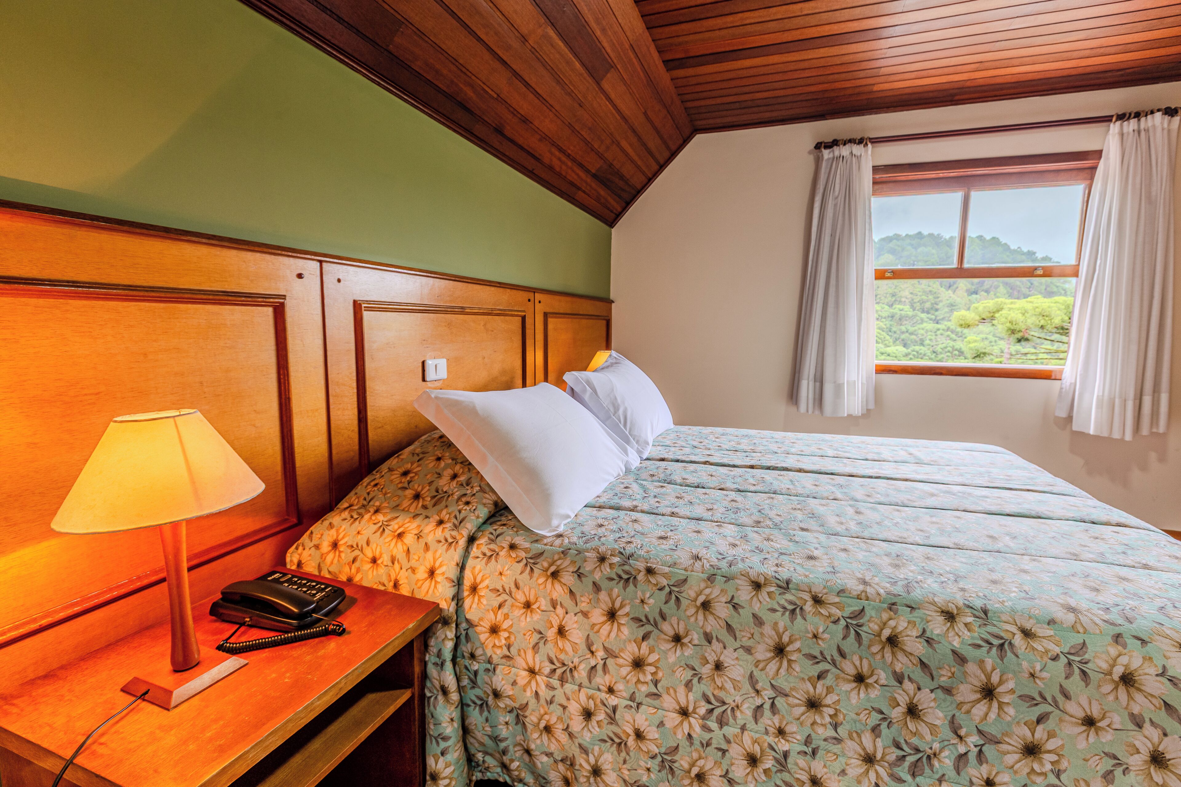 Una habitación de hotel con cabecera de madera, cama con ropa de cama floral, lámpara de noche, teléfono y ventana con cortinas.
